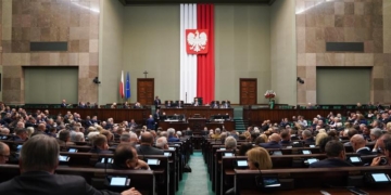 Archive photograph of the Sejm parliamentary chamber in Warsaw, with MPs seated beneath the Polish national emblem.