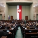 Archive photograph of the Sejm parliamentary chamber in Warsaw, with MPs seated beneath the Polish national emblem.