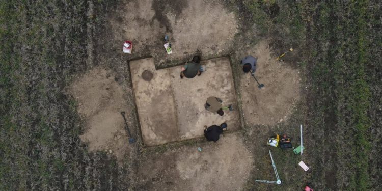 Overhead photograph of four archaeological researchers examining an excavation trench in a field at Gródek, eastern Poland. Image from the Stanisław Staszic Museum in Hrubieszów.