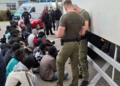 Border Guard personnel standing beside a refrigerated lorry while a group of people sit nearby with faces blurred.
