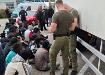 Border Guard personnel standing beside a refrigerated lorry while a group of people sit nearby with faces blurred.