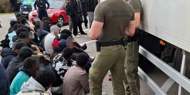Border Guard personnel standing beside a refrigerated lorry while a group of people sit nearby with faces blurred.