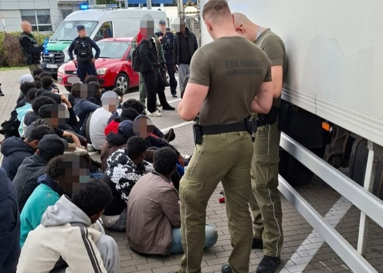 Border Guard personnel standing beside a refrigerated lorry while a group of people sit nearby with faces blurred.