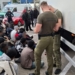 Border Guard personnel standing beside a refrigerated lorry while a group of people sit nearby with faces blurred.