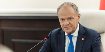 Government official seated at a meeting table with microphone, wearing a dark suit and blue tie.