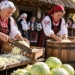 Editorial illustration of people chopping and pressing cabbage into a wooden barrel outside a rustic Polish farmhouse, with traditional-inspired dress and baskets of cabbages.