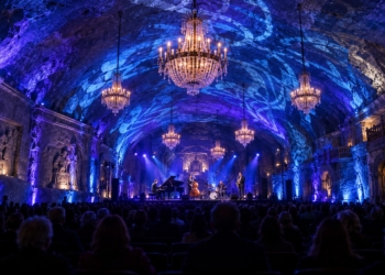 AI-generated editorial illustration of a jazz ensemble performing in an underground salt chamber illuminated with blue lighting and chandeliers, with an audience in the foreground