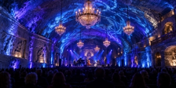 AI-generated editorial illustration of a jazz ensemble performing in an underground salt chamber illuminated with blue lighting and chandeliers, with an audience in the foreground