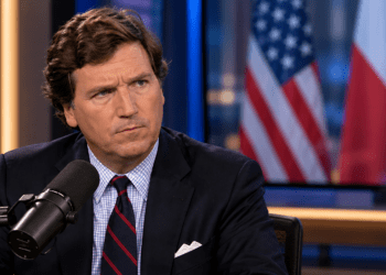 Tucker Carlson in a broadcast studio with blurred US and Polish flags on a background screen