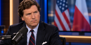 Tucker Carlson in a broadcast studio with blurred US and Polish flags on a background screen