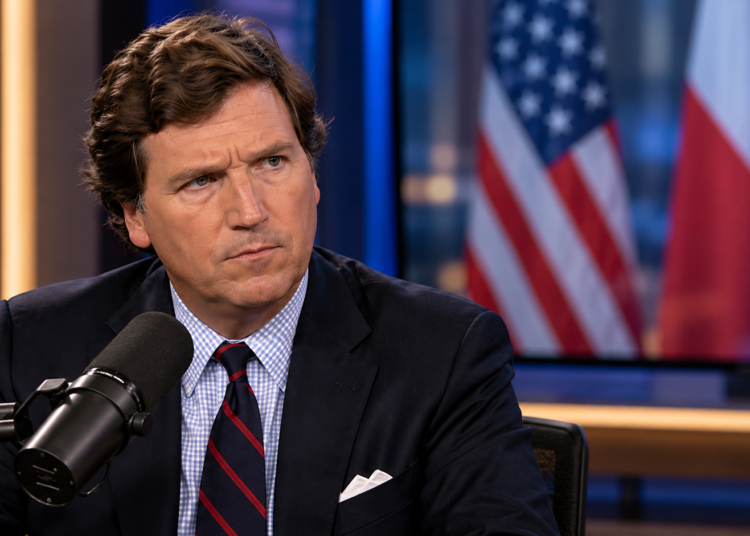 Tucker Carlson in a broadcast studio with blurred US and Polish flags on a background screen