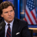 Tucker Carlson in a broadcast studio with blurred US and Polish flags on a background screen