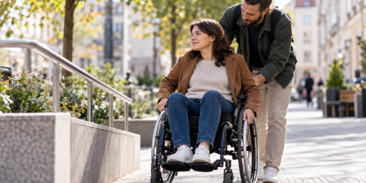 Digitally created image of a personal assistant helping a wheelchair user along a city pavement.