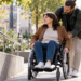 Digitally created image of a personal assistant helping a wheelchair user along a city pavement.
