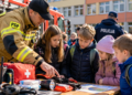 Digital illustration of a firefighter teaching children about emergency supplies outside a school, with fire and police personnel in the background.