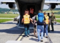 People accompanied by officers walking towards the open rear ramp of a military transport aircraft.