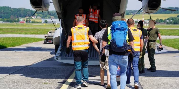 People accompanied by officers walking towards the open rear ramp of a military transport aircraft.
