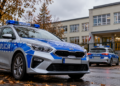 Digitally created editorial illustration showing two Polish police patrol cars parked outside a modern school building in autumn