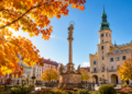 Digitally generated editorial illustration of golden autumn trees and a sunny historic Polish town square, representing unusually mild October weather.