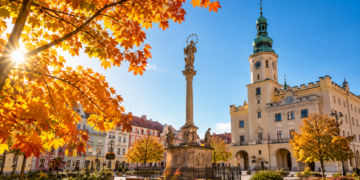 Digitally generated editorial illustration of golden autumn trees and a sunny historic Polish town square, representing unusually mild October weather.