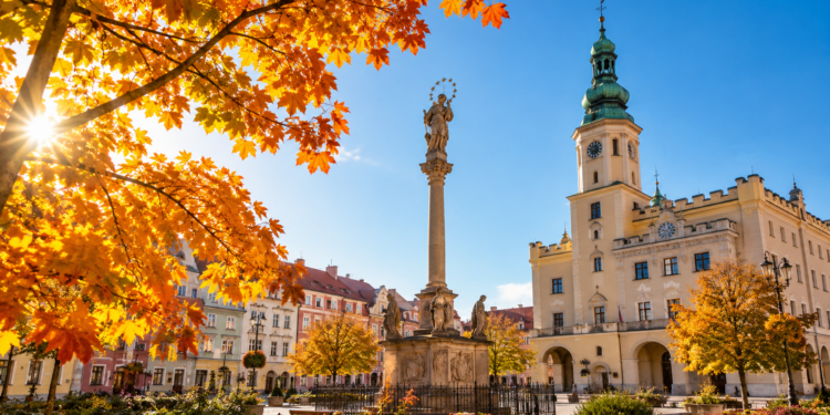 Digitally generated editorial illustration of golden autumn trees and a sunny historic Polish town square, representing unusually mild October weather.