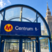 Digitally created illustration of Warsaw Centrum metro station's blue entrance sign and the Palace of Culture and Science behind it under a bright sky.